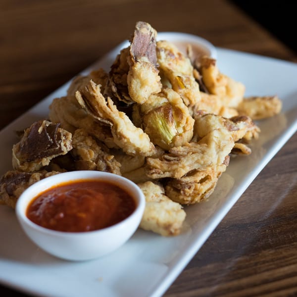Crispy fried artichoke hearts with a side of red dipping sauce on a white rectangular plate.