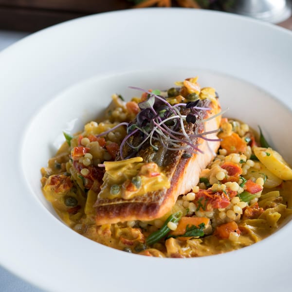 Pan-seared salmon with microgreens, served over couscous, vegetables, and a creamy sauce in a white bowl.