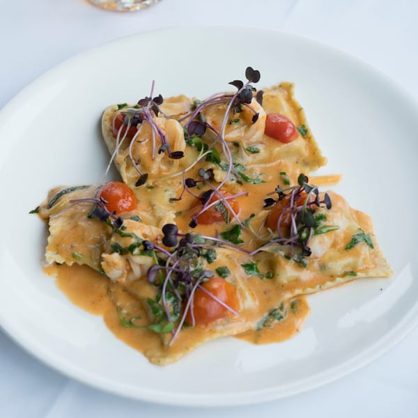 Dolce Restaurant's ravioli with creamy sauce, cherry tomatoes, shrimp, and microgreens on a white plate.