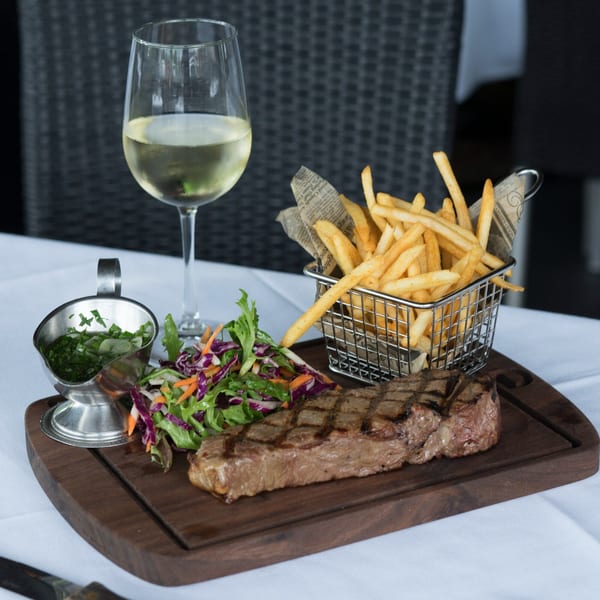Grilled steak, fries in a metal basket, salad, and a glass of white wine on a wooden board.