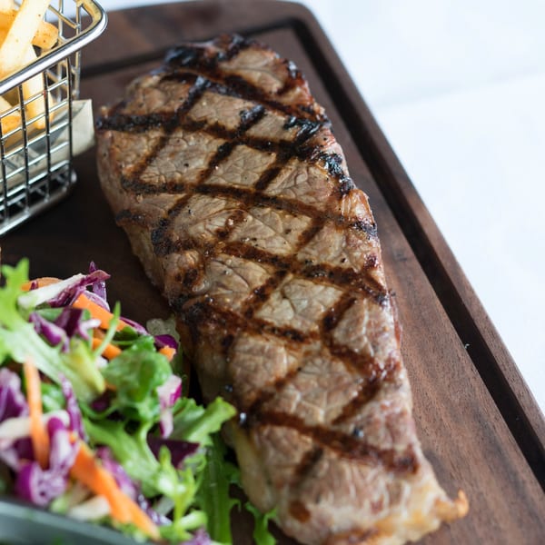 Grilled steak with crisscross sear marks, a side salad, and a basket of french fries on a wooden board.